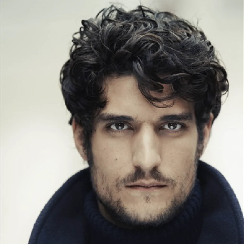 Primo piano di Louis Garrel, regista e attore protagonista del film L’Innocente (2022). Con uno sguardo intenso e capelli mossi, indossa un dolcevita scuro su sfondo chiaro, creando un contrasto elegante. L’immagine cattura il suo stile distintivo e la sua espressione magnetica.