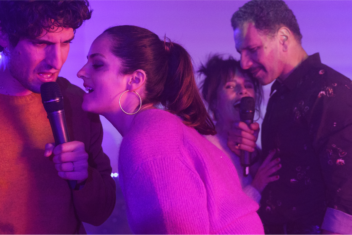 Scena festosa dal film L’Innocente (2022). Abel (Louis Garrel) e Clémence (Noémie Merlant) cantano con entusiasmo al karaoke, avvicinandosi con complicità ai microfoni. Sullo sfondo, Michel (Roschdy Zem) e Sylvie (Anouk Grinberg) si uniscono al divertimento, cantando e ballando. L’atmosfera è avvolta da luci viola e rosa, creando un’ambientazione vivace e spensierata.