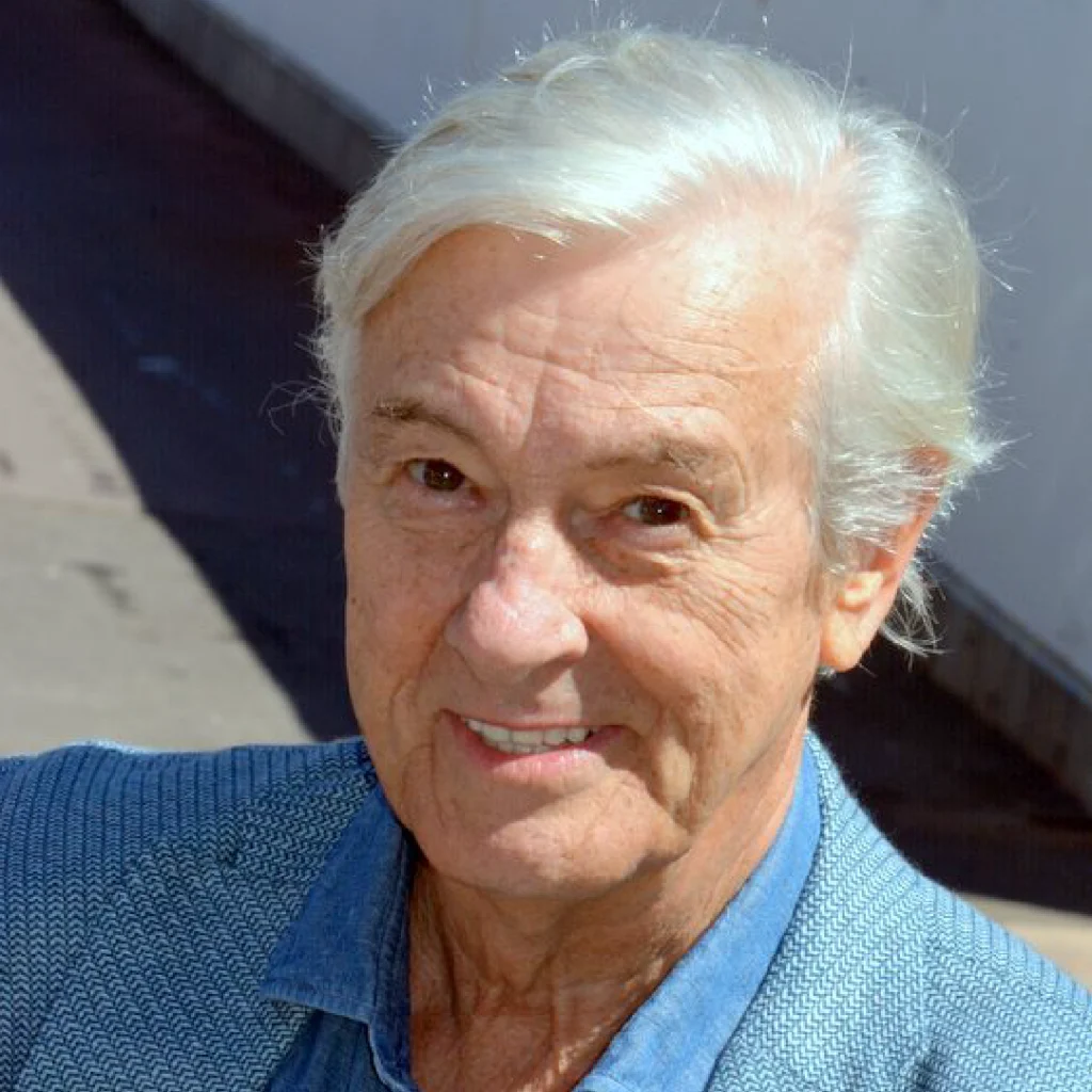 Primo piano del regista Paul Verhoeven con capelli bianchi e un sorriso accennato, vestito con una giacca blu chiaro.