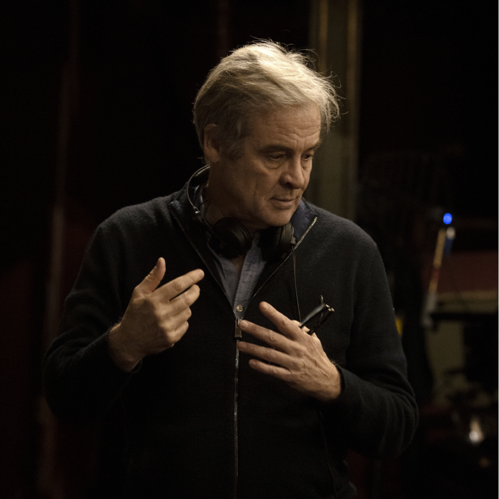 Il regista Emmanuel Courcol in una pausa sul set di L'Orchestra Stonata. Indossa un maglione scuro e delle cuffie intorno al collo mentre parla con espressione concentrata, indicando con le mani. Lo sfondo sfocato suggerisce l'ambiente di una sala prove o un set cinematografico.