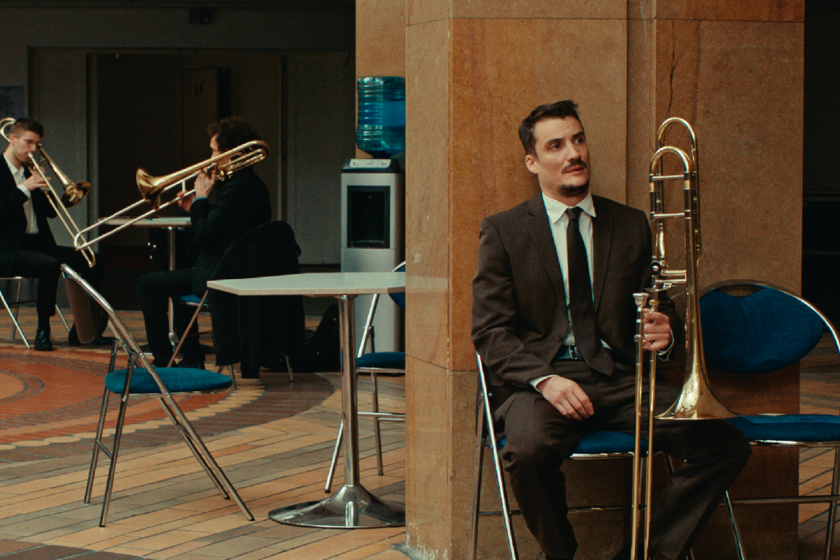 Pierre Lottin interpreta Jimmy, seduto con il suo trombone in un momento riflessivo del film L'Orchestra Stonata. Sullo sfondo, altri musicisti provano con i loro strumenti in un'area comune.