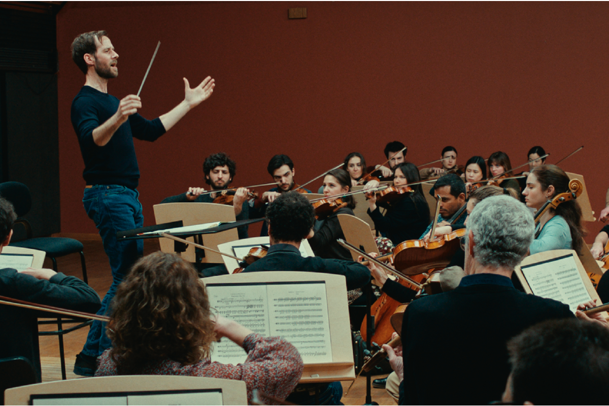 Scena dal film L'Orchestra Stonata: un direttore d’orchestra con bacchetta alza le braccia mentre guida un gruppo di musicisti intenti a suonare strumenti ad arco in una sala prove, creando un’atmosfera intensa e coinvolgente.