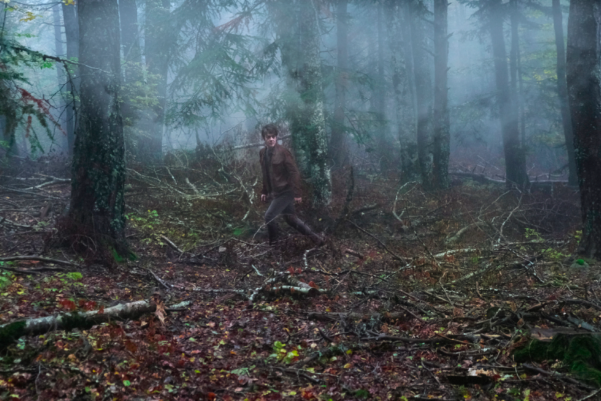 Scena tratta dal film “L’Uomo nel Bosco”. Jérémie (Félix Kysyl) cammina con un’espressione preoccupata attraverso una foresta fitta e oscura, avvolta dalla nebbia. I colori autunnali e il terreno ricoperto di rami spezzati creano un’atmosfera di mistero e tensione.