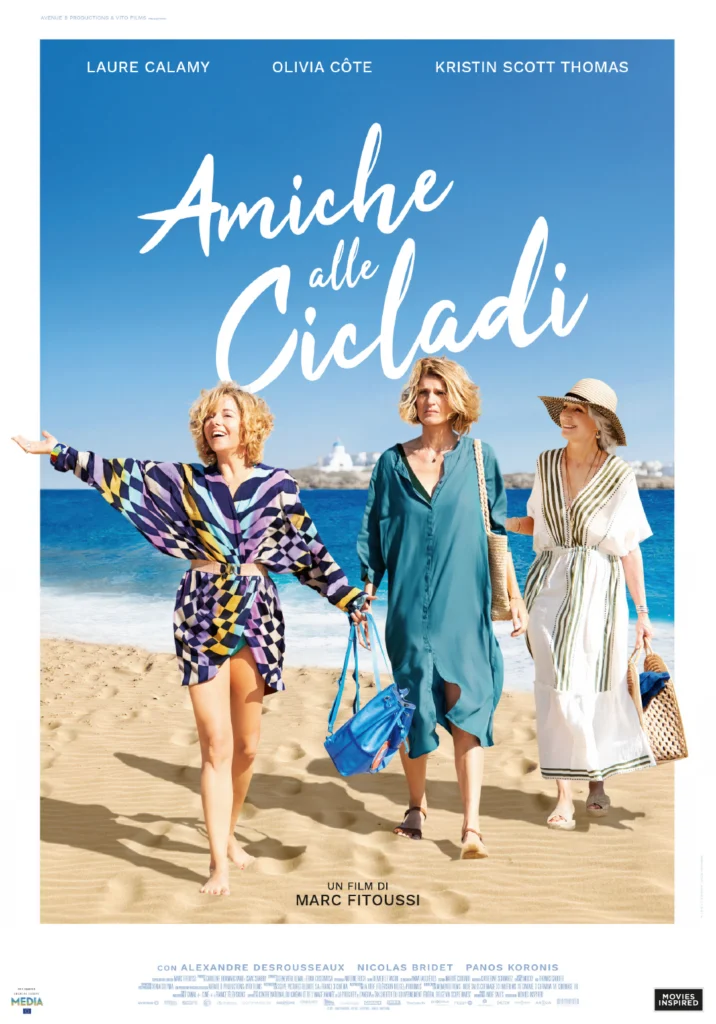 Le tre protagoniste di “Amiche alle Cicladi” camminano su una spiaggia dorata con il mare azzurro sullo sfondo. Laure Calamy sorride con un vestito colorato, Olivia Côte ha un’aria più seria in un abito blu, mentre Kristin Scott Thomas indossa un cappello di paglia e un abito bianco a righe.