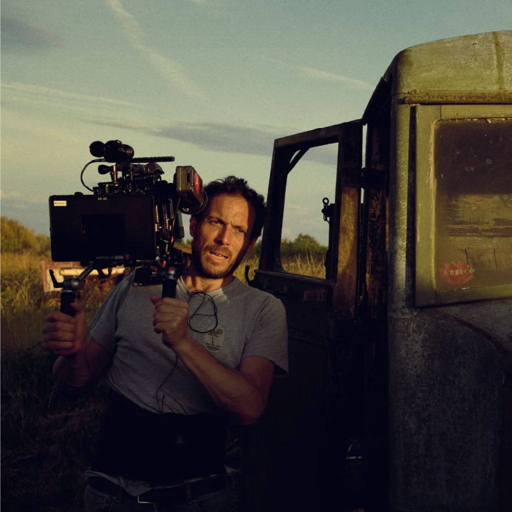 Il regista Olivier Casas mentre filma una scena con una cinepresa a spalla, accanto a un veicolo abbandonato in un paesaggio rurale.