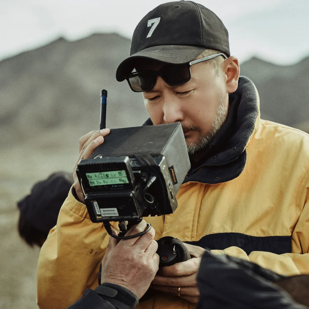 l regista Guan Hu con cappello nero e occhiali da sole mentre guarda nel mirino della cinepresa, sul set del film.